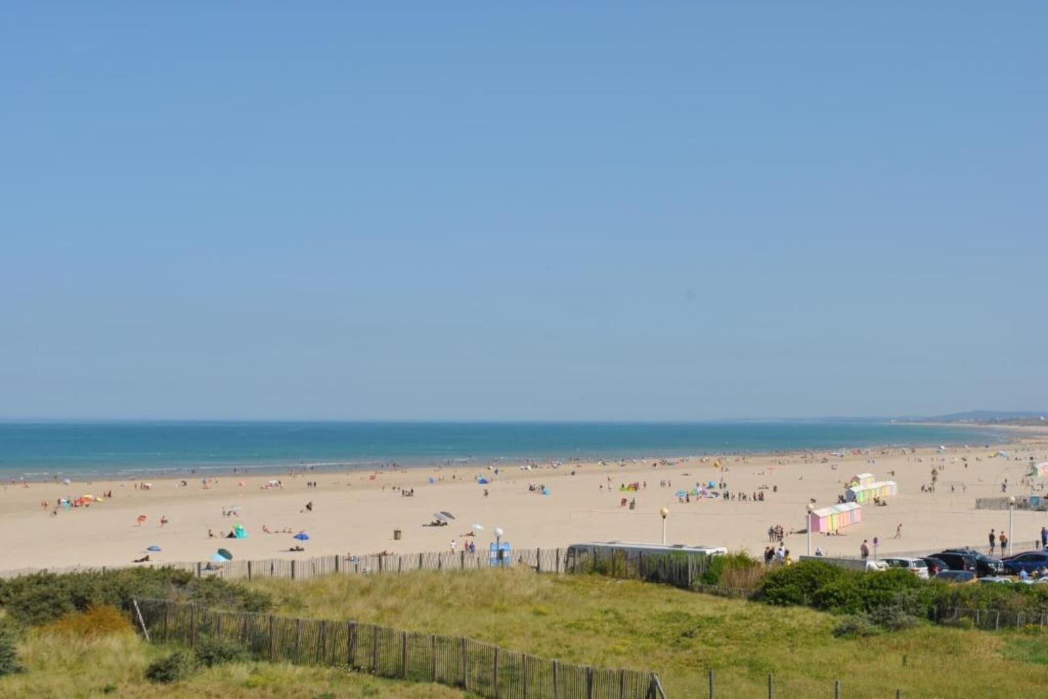 A beach with a wooden fence in the foreground, a clear sky, and the ocean in the background.