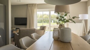 Un comedor con una mesa de madera, sillas y una lámpara grande. Hay un televisor montado en la pared y una ventana con vistas a zonas verdes.