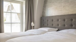Un dormitorio con dos camas, cabecero gris, lámpara de noche y ventana con cortinas.