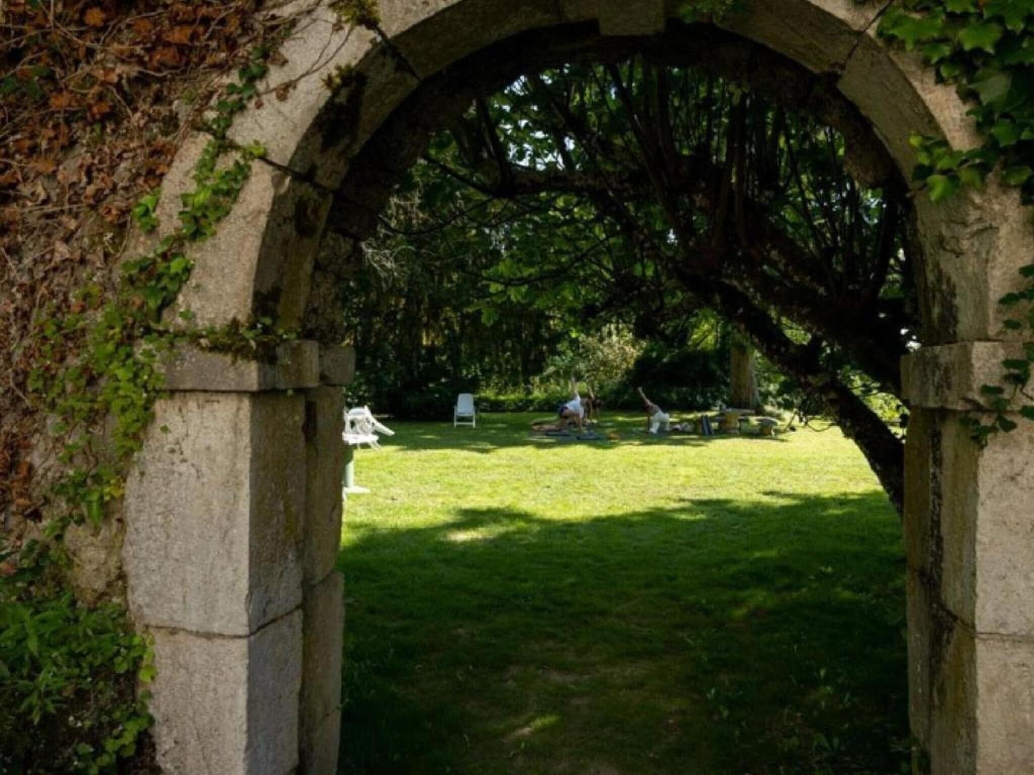 An arched stone structure with ivy, leading to a grassy area with two white chairs and a tree.