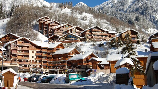 Un resort de montaña cubierto de nieve con edificios de madera, un estacionamiento y una vista despejada de las montañas circundantes.