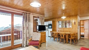 A wooden-floored room with a dining table, chairs, a TV, and a balcony with curtains. - Magnificent chalet with balcony (Champagny en Vanoise)