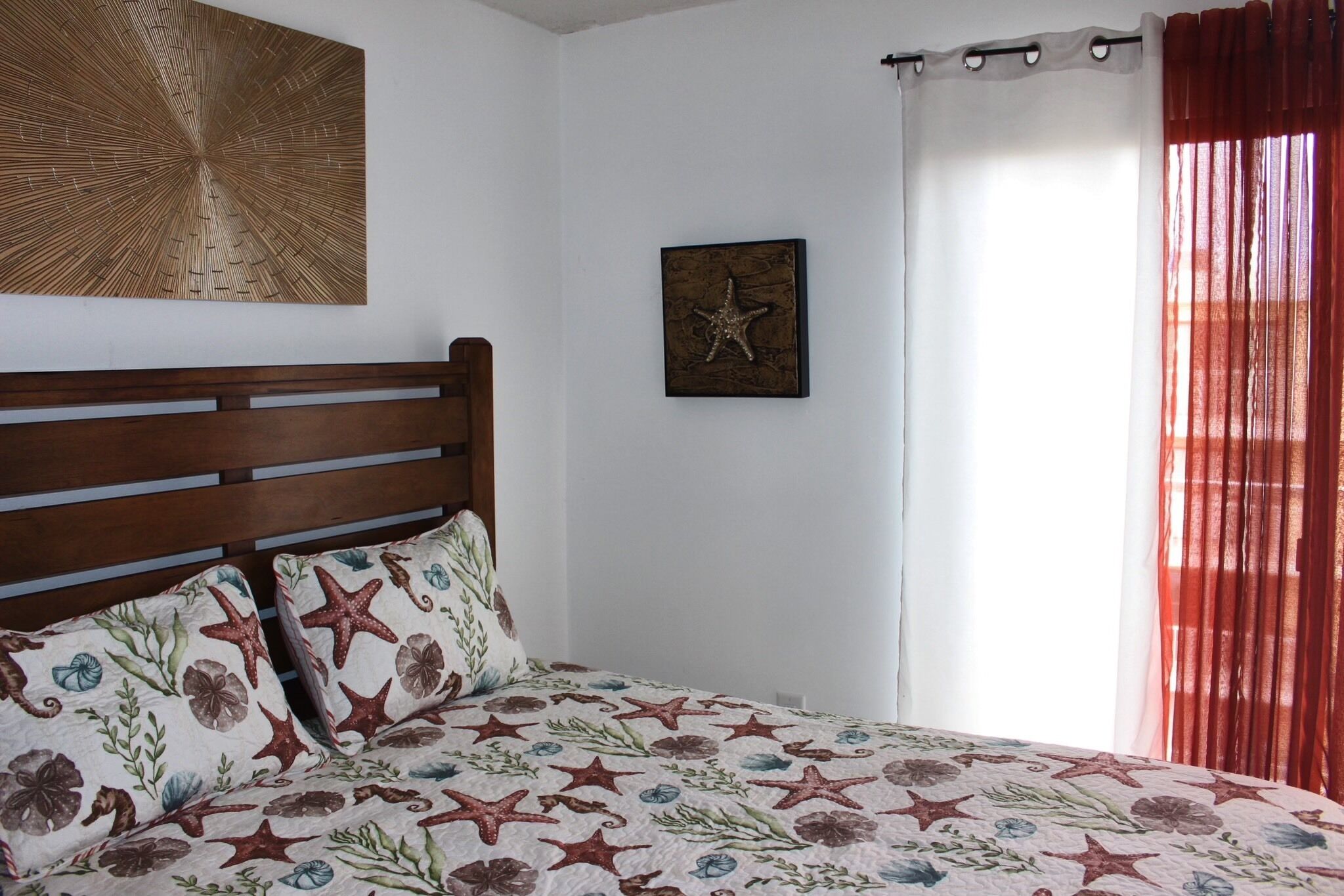 Ein Schlafzimmer mit einem Holzkopfteil, einem Bett mit Tagesdecke mit floralem und Sternmuster, einem Wandbild und einem Fenster mit roten Vorhängen.