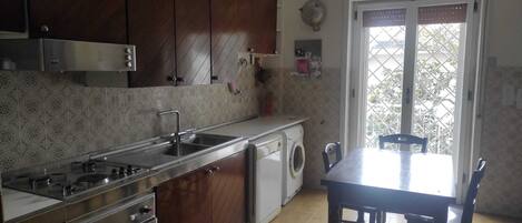 A compact kitchen with a sink, stove, oven, and a washing machine. There is a dining table with chairs and a window with a view of greenery outside.