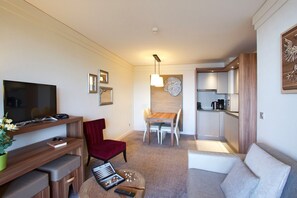 A modern living room with a flat-screen TV, a red chair, a dining table with chairs, and a kitchen area with cabinets and appliances.