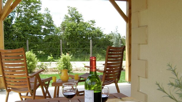 Een buitenterras met houten stoelen en tafels, een fles wijn en een bord met eten.
