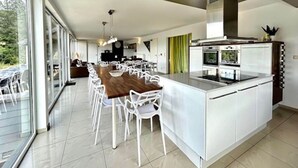 A modern kitchen with white cabinetry, a large island with a built-in oven, and a dining area with white chairs. - Villa in Coo with Pool & Panoramic Views (Coo - Stavelot)
