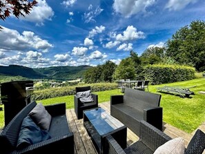 Un área de estar al aire libre con mobiliario de mimbre, una mesa y vistas a un paisaje exuberante con colinas y árboles.