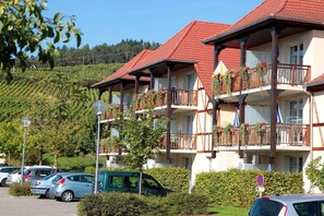 Un hotel de varios pisos con balcones, un estacionamiento y un viñedo al fondo.