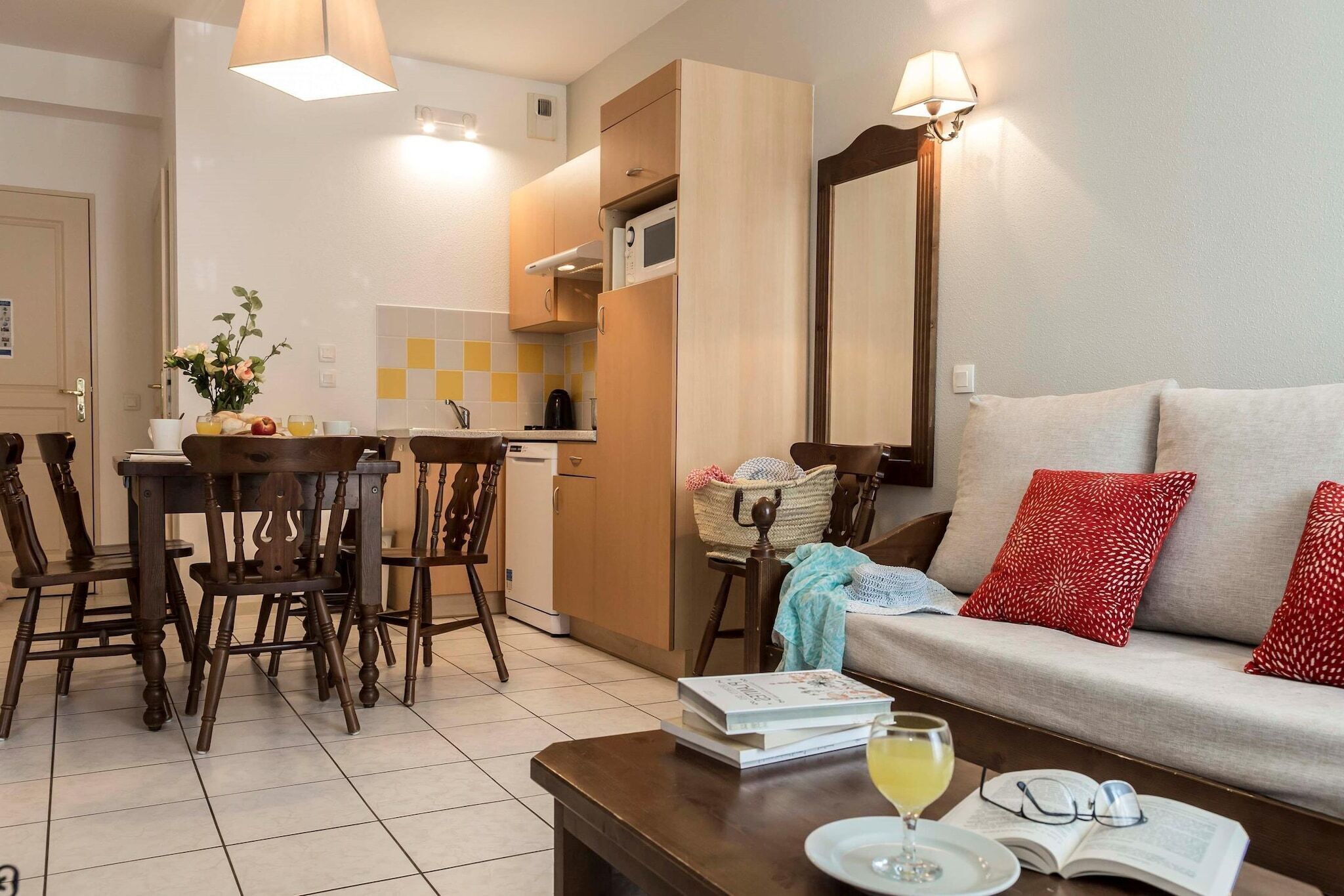 A living room with a sofa, a coffee table with books and a glass of wine, a dining area with chairs and a table, and a kitchen area with a microwave.