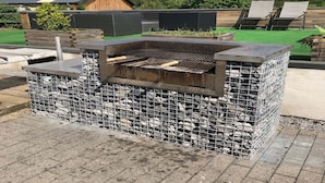 An outdoor grill with a stone and metal design, surrounded by a patio area with chairs and a wooden structure in the background. - House in Moulin du Ruy with Forest Views (Stoumont-Moulin du ruy)