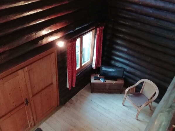 Interior de una cabaña con una puerta de madera, una ventana con cortinas rojas, un televisor sobre una mesa de madera y una silla.