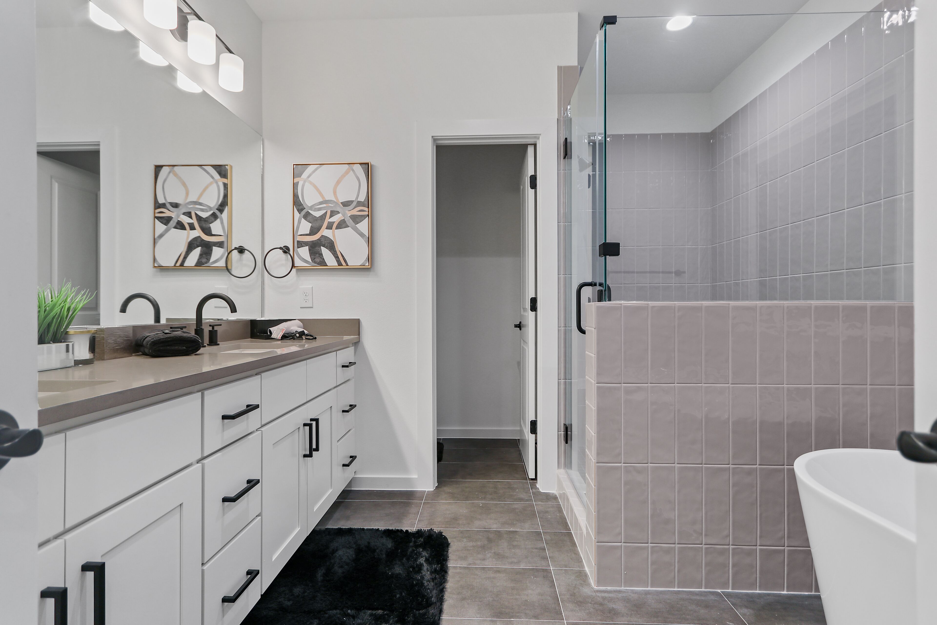 A modern bathroom with a glass shower enclosure, a white sink with a beige countertop, and two abstract paintings on the wall.