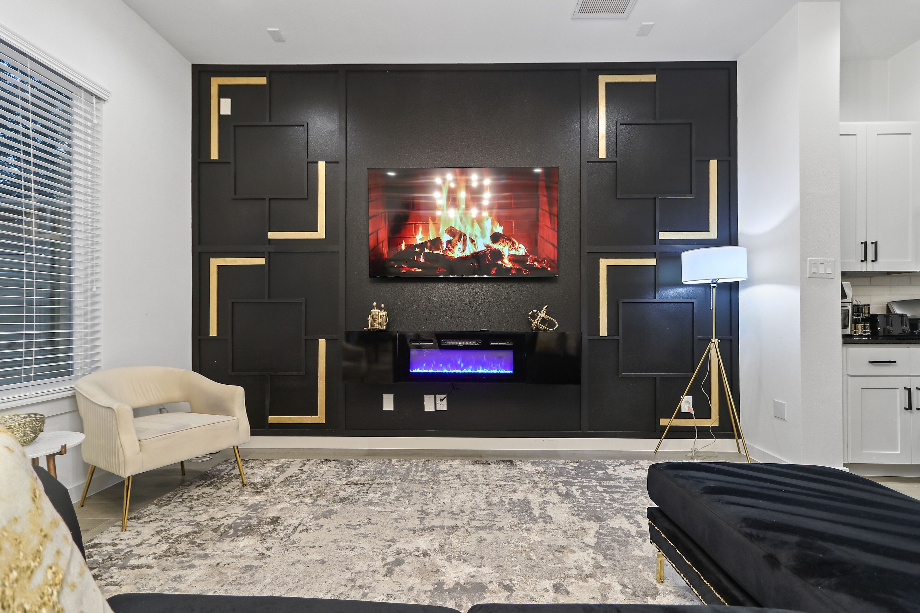 Un salon moderne avec un grand téléviseur à écran plat fixé sur un mur noir orné de motifs géométriques, un fauteuil beige, un canapé noir et une petite table d’appoint.