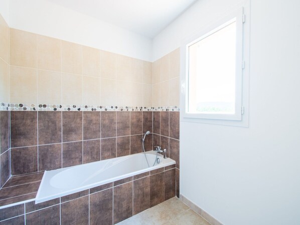 A bathroom with a bathtub, beige and brown tiles, a window, and a shower.