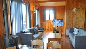 Et hyggeligt sommerhusinteriør med sofa, spisebord med stole, fjernsyn og en brændeovn.