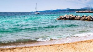 A beach with clear turquoise water, a rocky breakwater, and sailboats in the distance.