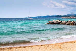 Ein Strand mit klarem türkisblauem Wasser, einem steinernen Wellenbrecher und Segelbooten in der Ferne.