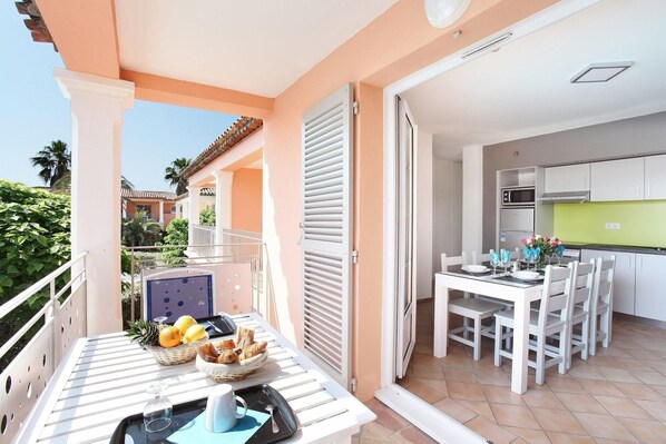 Un balcón con una mesa dispuesta para el desayuno, una cocina con armarios blancos y vista a una palmera.