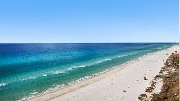A beach with clear blue water, sandy shore, and a pier in the distance.