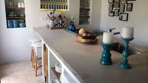 Eine Küche mit einer zentralen Kochinsel, weißen Barhocker, einer Obstschale und einem an der Wand befestigten Regal mit Küchenutensilien.