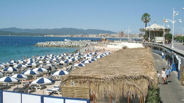 Ein Strand mit vielen Sonnenschirmen und Liegestühlen, einer Struktur mit Reetdach und einer Promenade mit einer Palme und einem Karussell.