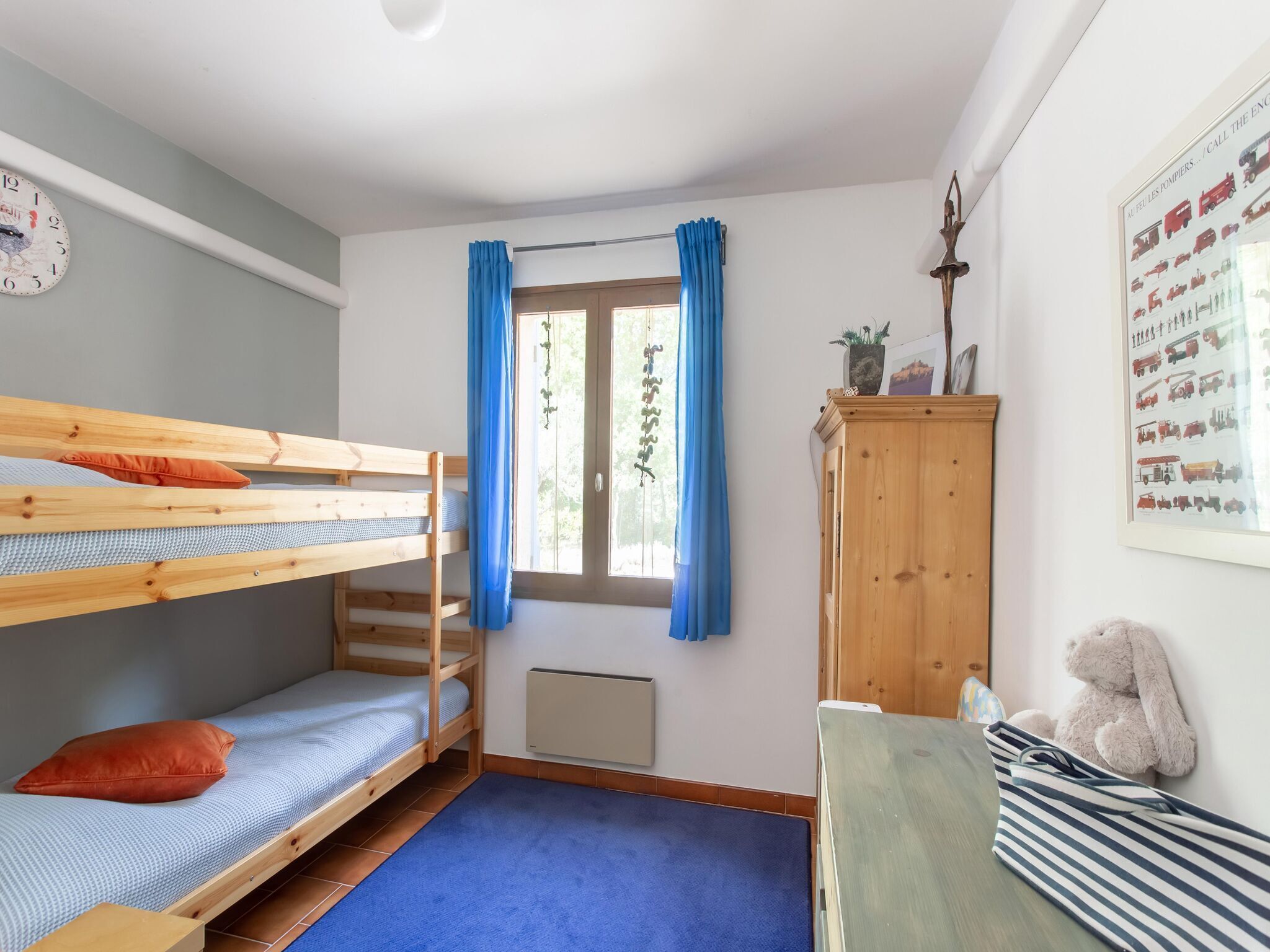 Ein Zimmer mit Etagenbetten, einem Heizkörper, einem Fenster mit Vorhängen, einem Holzschrank und einem Teddybären.