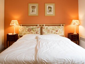 Una cama bien hecha con sábanas blancas en una habitación con paredes anaranjadas, dos cuadros en la pared y dos mesitas de noche con lámparas.