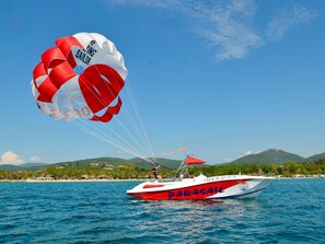 Un bote de parasailing llamado 'PARASAIL' con diseño rojo y blanco, anclado en aguas tranquilas con un paisaje montañoso al fondo.
