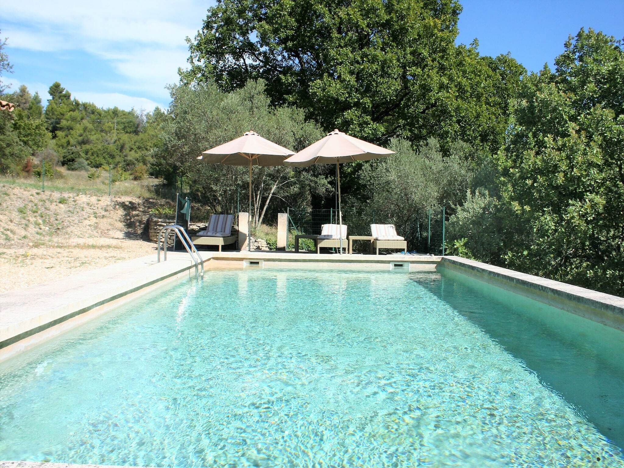 A swimming pool with clear water, surrounded by lounge chairs and umbrellas, set against a backdrop of trees and a clear sky.