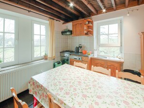 A cozy kitchen with wooden cabinets, a dining table with a floral tablecloth, and a view of the outdoors through large windows. - Spacious Farmhouse Retreat (Brisy-Gouvy)