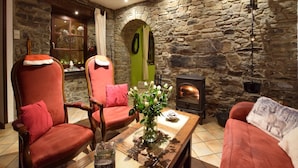 Une pièce chaleureuse aux murs de pierre, avec une cheminée, deux fauteuils en cuir rouge, une table en bois ornée d’un vase de fleurs et une porte verte.