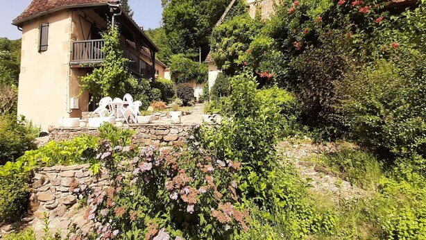A traditional stone house with a balcony, surrounded by lush greenery and flowering plants. - Charming 1930s Home in Altillac (Altillac)