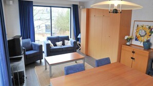 A modern living room with a sofa, chairs, a coffee table, and a TV. There is a large window with curtains, a wooden cabinet, and a painting on the wall. - Villa Apartment with Terrace (Durbuy)