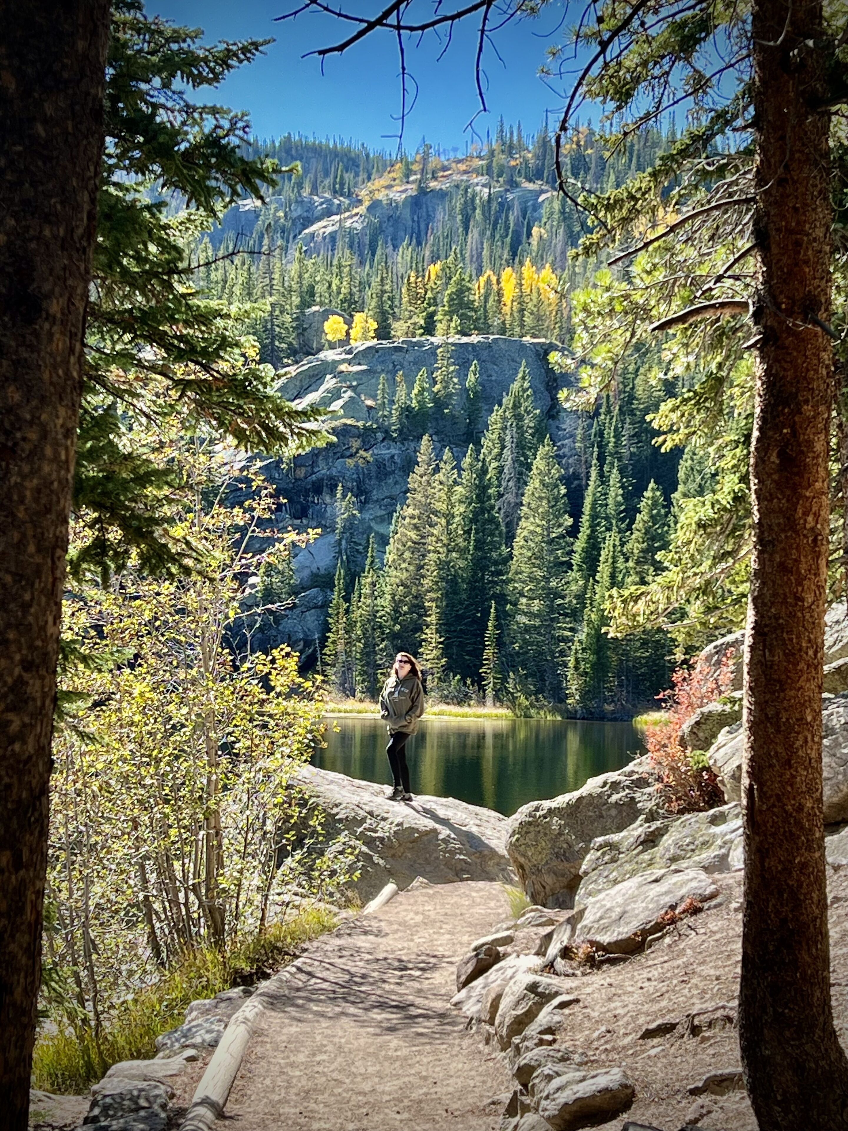 Une personne se tient sur un sentier rocheux, avec un lac et une forêt en arrière-plan.