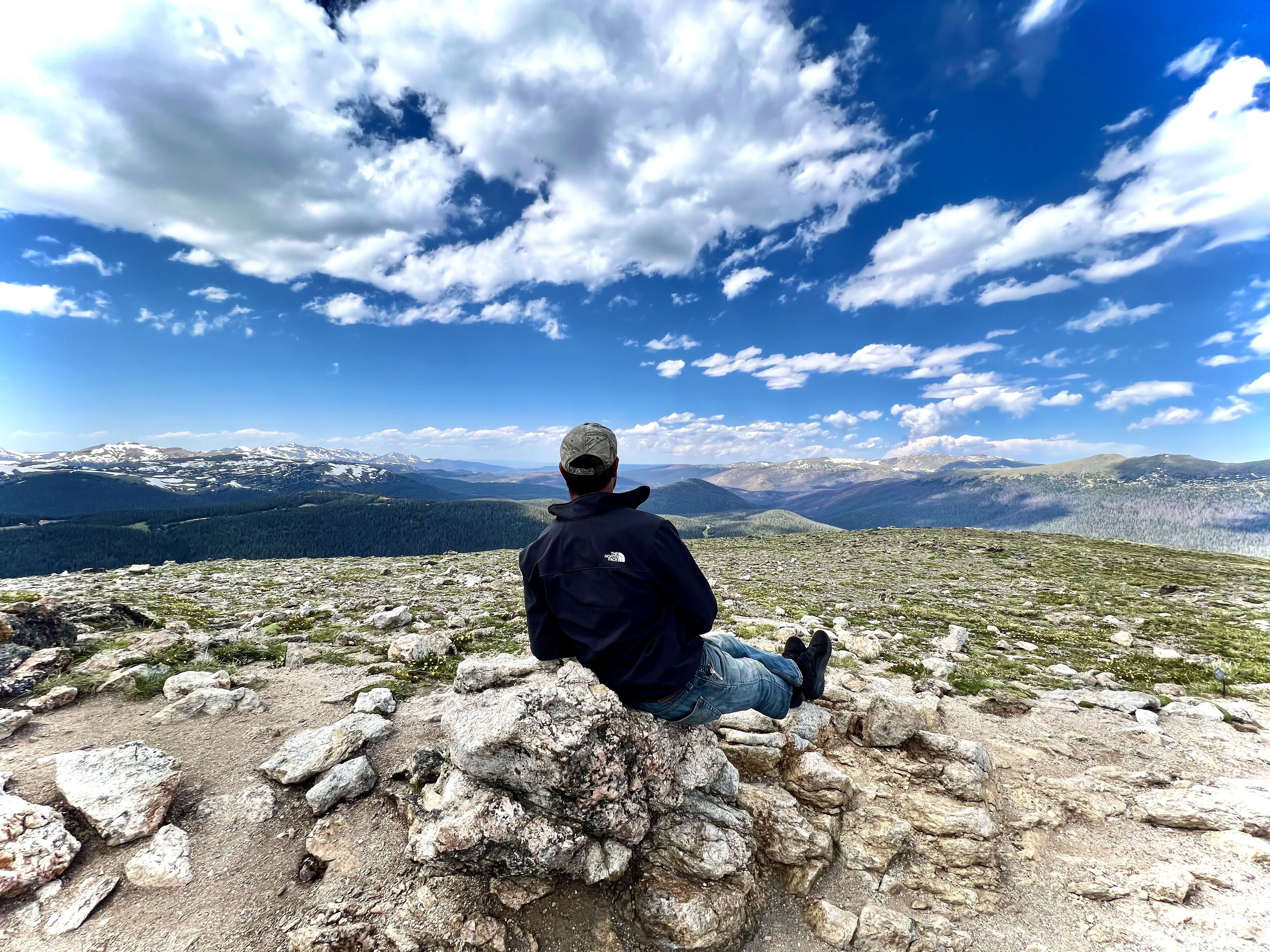 Une personne assise sur un promontoire rocheux, d’où elle domine un paysage montagneux sous un ciel dégagé.