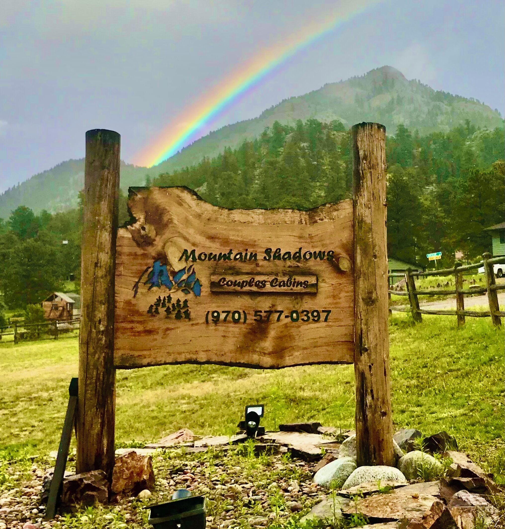 A wooden sign with 'Mountain Shadows' and a phone number, set against a mountain backdrop with a rainbow.