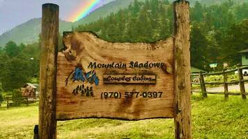 A wooden sign with 'Mountain Shadows' and a phone number, set against a mountain backdrop with a rainbow.