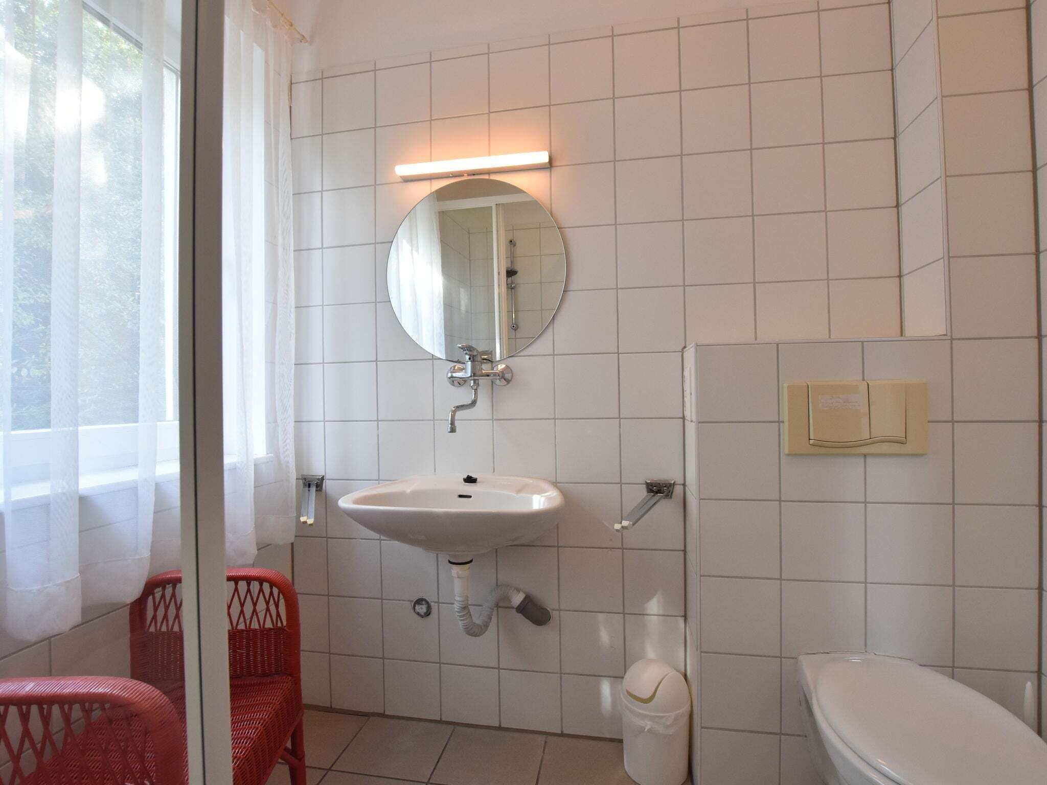 Ett badrum med ett vitt handfat, en spegel och en toalett. Det finns ett fönster med tunna, genomskinliga gardiner och en röd stol i hörnet.