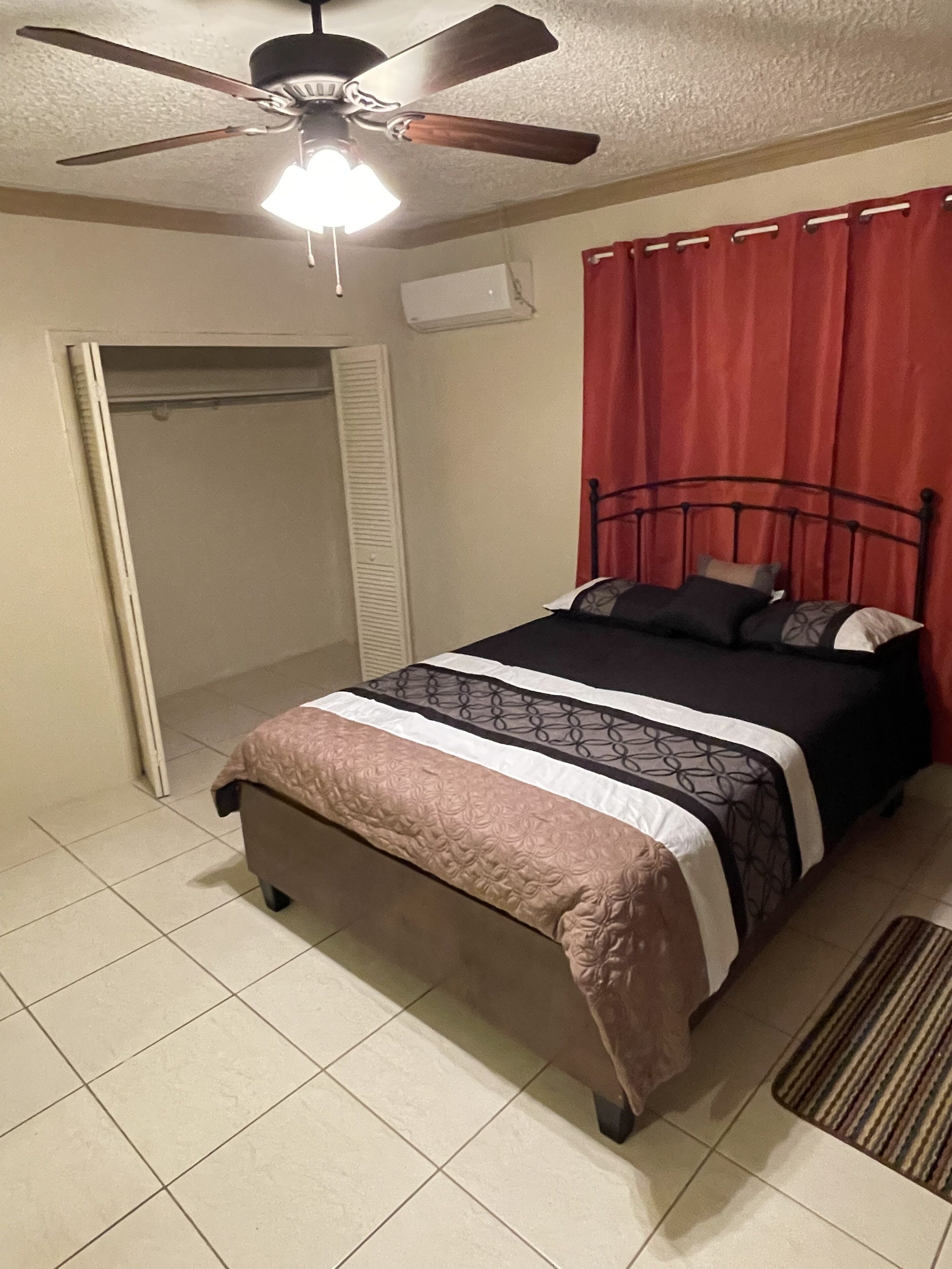 A bedroom with a bed, a ceiling fan, a window with red curtains, and a door with a white shutter.