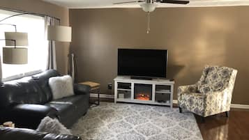 A living room with a leather sofa, a patterned armchair, a TV on a white entertainment unit, and a ceiling fan.