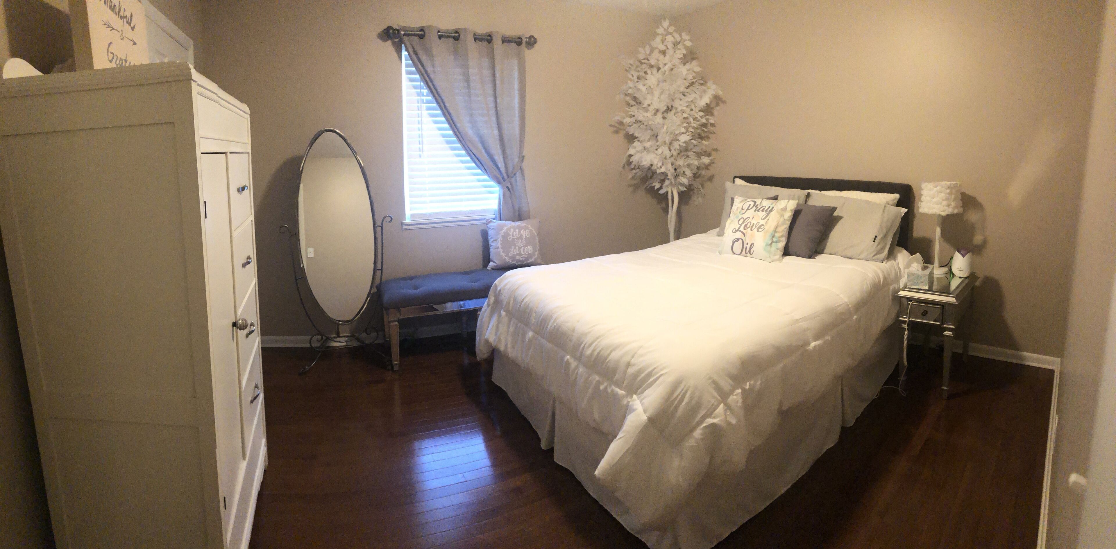 A bedroom with a bed, a dresser, a vanity with a mirror, a window with curtains, and a nightstand with a lamp.