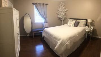 A bedroom with a bed, a dresser, a vanity with a mirror, a window with curtains, and a nightstand with a lamp.