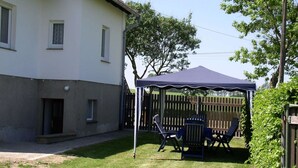 A covered outdoor seating area with a table and chairs, a wooden fence, and a house in the background. - Charmantes Apartment in Jennewitz, in Ostseenähe (Jennewitz)