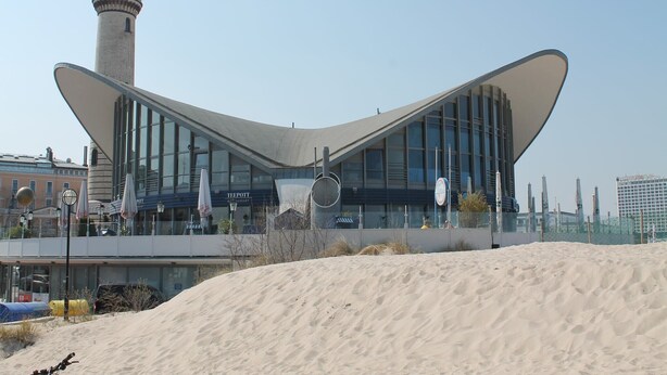 A modern building with a unique curved roof design, a prominent tower, and a sandy foreground. - Charmantes Apartment in Jennewitz, in Ostseenähe (Jennewitz)
