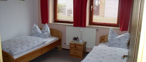 A bedroom with two wooden beds, a small bedside table, a radiator, and two windows with red curtains.