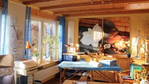 A room with wooden beams, a massage table, a window with plants, a statue, and a painting of a flower.