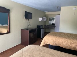 Habitación de hotel con dos camas, un escritorio con computadora, un televisor montado en la pared y una pequeña kitchenette.