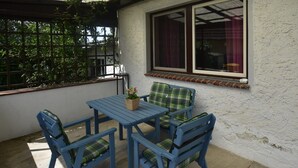 A covered patio area with a table and four chairs, a window with curtains, and a view of greenery outside. - Ferienwohnung auf dem Land in Strandnähe (Kröpelin / OT Diedrichshagen)