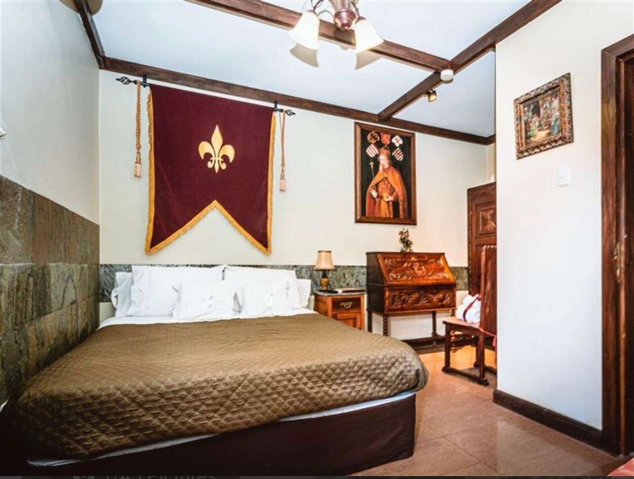 A bedroom with a large bed, a wooden nightstand, a desk, and a chair. There is a red banner with a fleur-de-lis symbol on the wall.